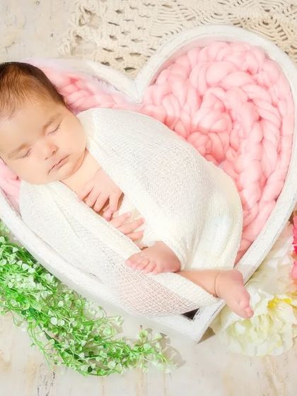 A newborn sleeps soundly in a heart-shaped basket, surrounded by soft greenery and flowers. This overhead shot is a beautiful composition of love and new life.