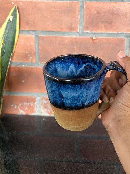 A hand-held view of a wheel-thrown mug with a stunning blue and black glaze that contrasts with the raw clay base.