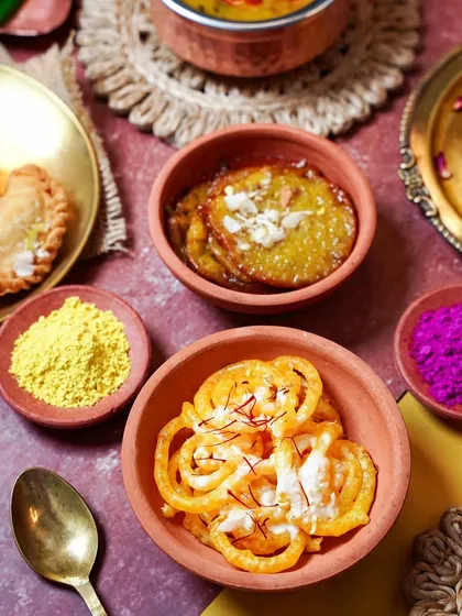 A close-up of classic Holi sweets, Jalebi and Malpua, served in terracotta bowls. The warm lighting and scattered colors evoke the sweet indulgence of the festival.