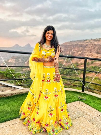 A bride in a stunning yellow lehenga, posing against a scenic mountain backdrop with her mehandi.