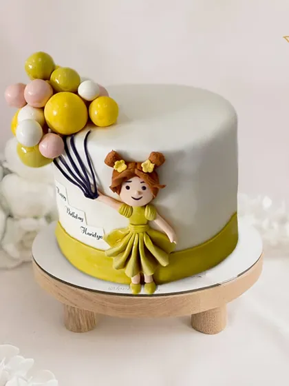 A charming and whimsical cake featuring a little girl holding a bouquet of balloons. The clean design and cute fondant figurine make this a sweet and modern choice for a child's birthday.