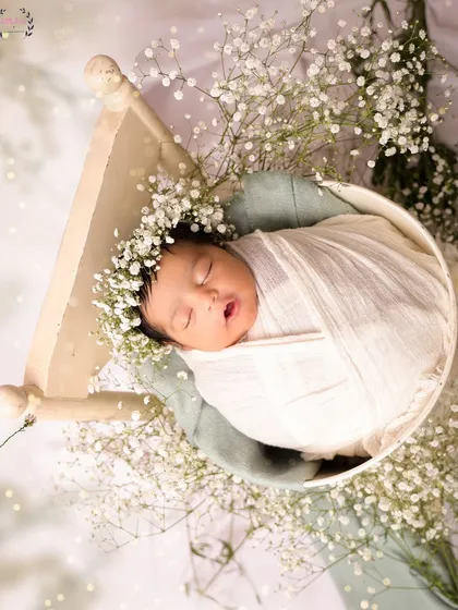 A newborn baby, wearing a floral crown, is swaddled in white and rests in a tiny bed surrounded by baby's breath. This overhead shot creates a beautiful, nest-like composition.
