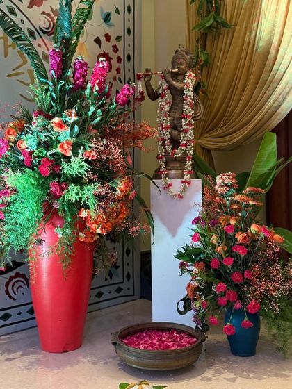 A detail of a traditional entrance, featuring a Krishna statue adorned with a floral garland. The space is decorated with vibrant floral arrangements in red and orange, and a brass 'urli' bowl with floating petals, infusing the arrival with culture and charm.