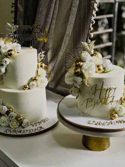 Two milestones, one elegant theme. This photo showcases a matching two-tier anniversary cake and a single-tier birthday cake, both adorned with white florals and luxe gold accents for a cohesive celebration.