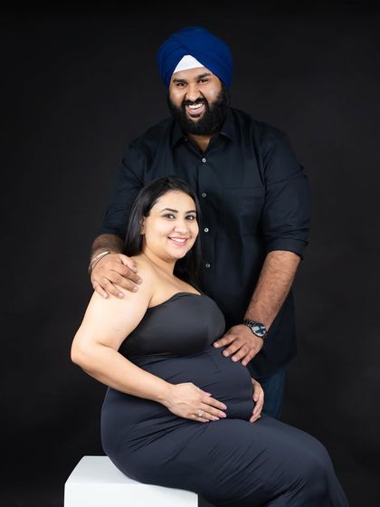 A joyful and classic studio portrait. This couple shares a happy moment, with the partner's arm around the mom-to-be, both smiling warmly for the camera.