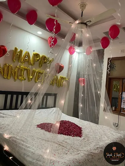 An anniversary room decoration featuring a fairy light canopy, "HAPPY ANNIVERSARY" foil balloons, red ceiling balloons, and a heart made of rose petals on the bed.