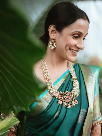 A close-up of a radiant bride, highlighting the intricate details of her engagement look. From the stunning pearl and gold necklace to the traditional nath and the subtle glow of her makeup, every element is chosen to complement her natural beauty and the rich teal of her saree.
