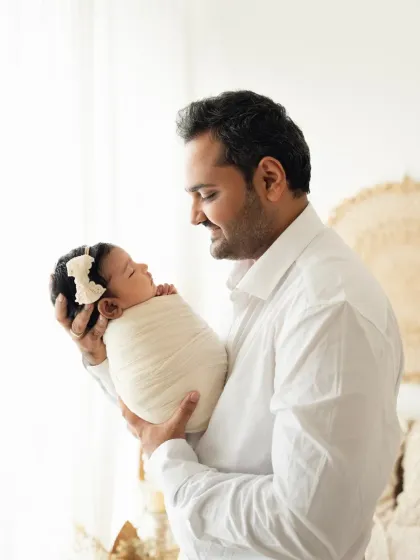 A proud father holds his sleeping newborn daughter, gazing at her with a smile. This portrait showcases the special bond between a father and his new baby.