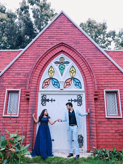 A romantic pre-wedding photo in front of a charming chapel-like building. The navy blue gown and light blue suit are a classic and elegant combination.