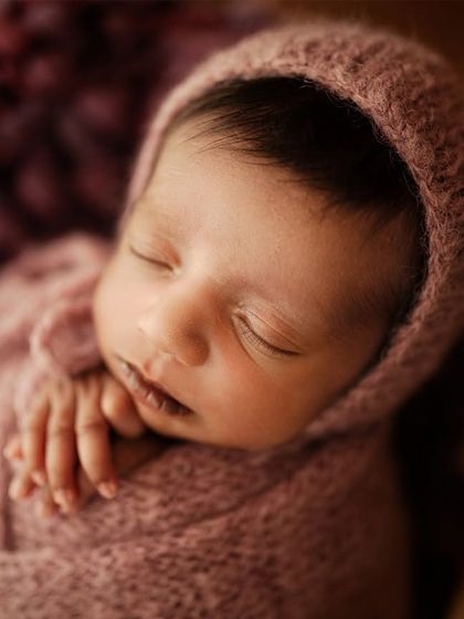 A detailed close-up of a newborn's face, highlighting their perfect features while they sleep. The soft pink wrap and bonnet add a gentle warmth.
