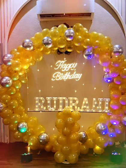 A bright and cheerful birthday setup with a yellow balloon ring, silver disco balls, a neon sign, and light-up letters for the name "Rudrani".