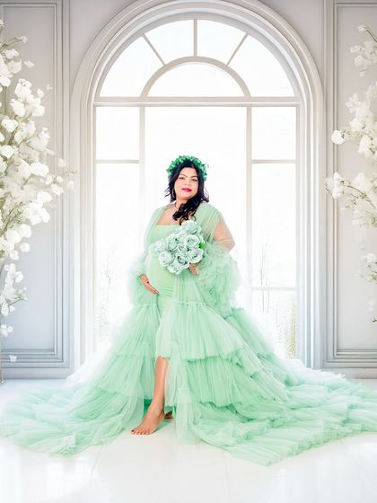 A full-length solo portrait of the mom-to-be in a beautiful mint green ruffled gown. The bright, airy backdrop with white arches and cherry blossoms creates a fresh and elegant look.
