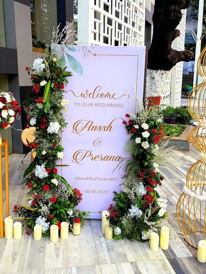 This welcome setup combines traditional florals with modern design. A simple, elegant sign is flanked by arrangements of red and white roses, while golden birdcage-like structures add a touch of contemporary artistry.