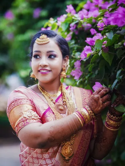 The bride poses gracefully amidst bougainvillea flowers, her traditional pink and gold saree creating a beautiful contrast.