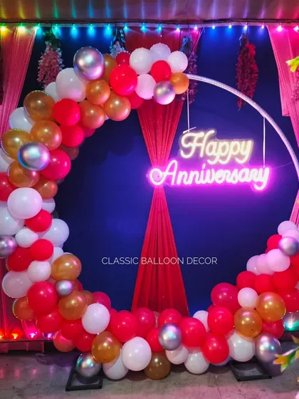 A vibrant anniversary ring decoration for a party. This setup uses red, white, and gold balloons against a red curtain backdrop, with a bright 'Happy Anniversary' neon sign.