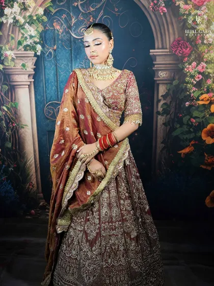 A full view of a classic bridal look, featuring a deep maroon lehenga with intricate gold work. The makeup is kept warm and glowing, with a focus on defined eyes and a neutral lip to let the outfit shine.