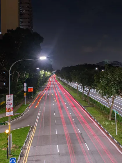 A long-exposure shot of a Singaporean road at night, with the red light trails of traffic creating straight lines into the distance.