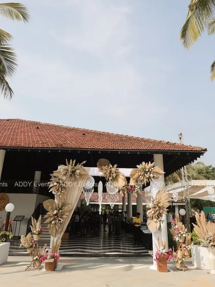 A grand entrance to a tropical-themed Mehndi, with arches decorated with pampas grass, dried leaves, and dreamcatchers set against a backdrop of palm trees.