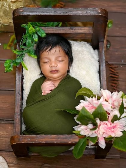 A newborn wrapped in green, sleeping peacefully in a small wooden crate. The surrounding flowers and greenery add a touch of nature to this studio setup.