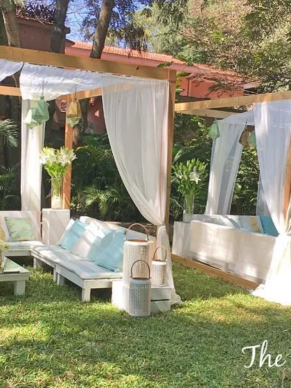 I created these custom-built wooden cabanas with flowing white drapes to provide a relaxed and breezy lounge area for an outdoor party. The space is furnished with comfortable seating, soft blue and white cushions, and rustic lanterns to establish a chic, beach-inspired vibe.
