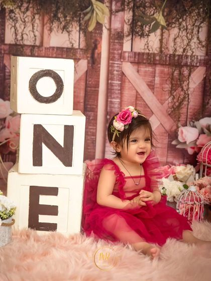 A happy one-year-old girl sitting next to "ONE" blocks in a beautiful floral and rustic barn-door setting.