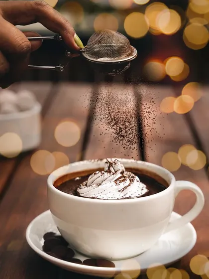 A classic cup of hot chocolate with whipped cream, with festive bokeh lights in the background. The action of dusting cocoa powder on top adds a final, delicious touch.
