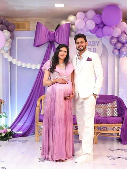 The happy couple posing for a photo at their bow-themed baby shower. The backdrop of purple and silver balloons creates a beautiful and celebratory setting.