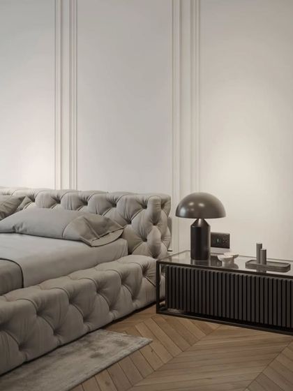A close-up of a luxurious tufted bed in a sophisticated grey fabric. The herringbone wood flooring and classic wall molding add to the room's timeless elegance.