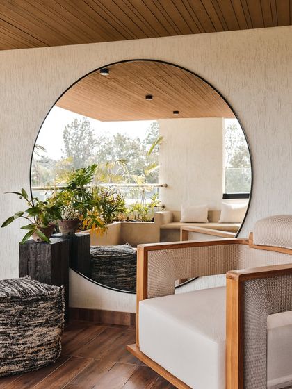 A large circular mirror in this semi-outdoor space reflects the lush greenery outside, blurring the boundary between indoors and out. The use of natural materials like wood and cane enhances the organic feel.