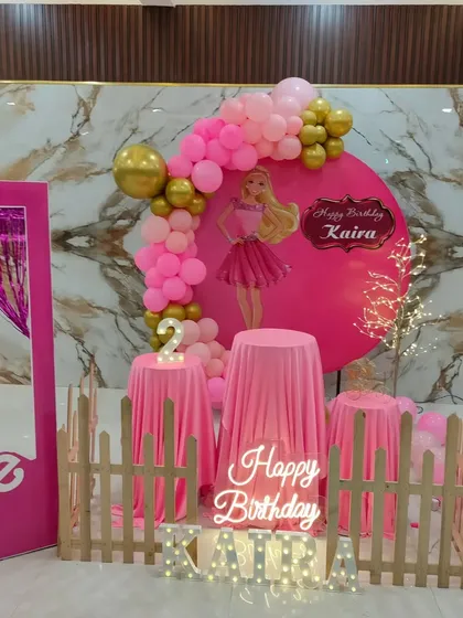 A stylish Barbie party setup against a marble-patterned wall, featuring a pink backdrop, balloon arch, and a fun photo box.