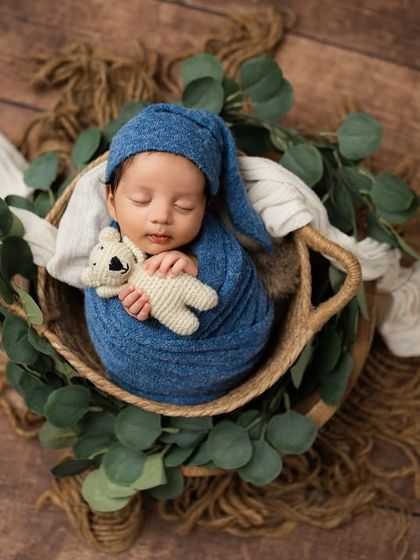 A wide shot showing the complete setup, with the baby and his teddy bear nestled in a basket surrounded by eucalyptus. This is a perfect example of a themed portrait.