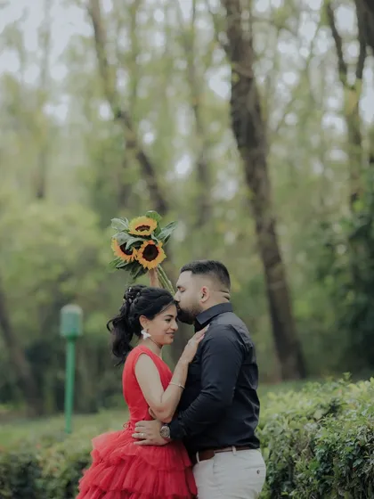 A tender moment where the groom kisses his bride-to-be's forehead. Her stunning red dress and the natural park setting create a beautiful contrast, making this a classic romantic portrait.