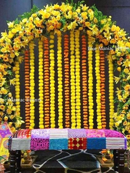 A unique Mehendi seating idea with a patchwork bench set against a backdrop of hanging marigold and gladiolus flower strings, framed by a floral arch.