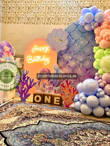 A "one"derful mermaid-themed first birthday with a neon sign and a rainbow pastel balloon garland. The iridescent scale backdrop adds a magical touch.