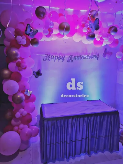 This anniversary party backdrop features a 'Happy Anniversary' sign against a simple white background, framed by a balloon arch in shades of pink and gold with butterfly accents. The purple uplighting adds a dramatic effect.