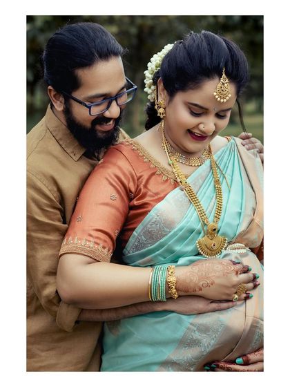 A close-up portrait of a couple during their Seemantham ceremony. The husband embraces his wife from behind as they both look down at her baby bump, sharing a moment of quiet joy.