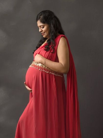 A profile shot capturing the serene glow of pregnancy. The rich red gown stands out beautifully against the dark, textured backdrop, emphasizing the beautiful silhouette of the baby bump.
