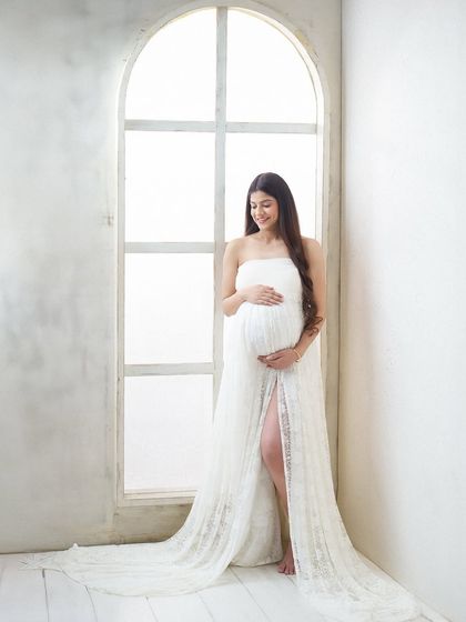 A smiling portrait of a mother-to-be in a strapless white lace gown, standing by a large, bright window. The natural light fills the scene, creating a joyful and airy feel.