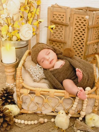 A sleeping newborn in a brown knit outfit, resting in a small wicker bed as part of a warm, earthy-toned photoshoot.