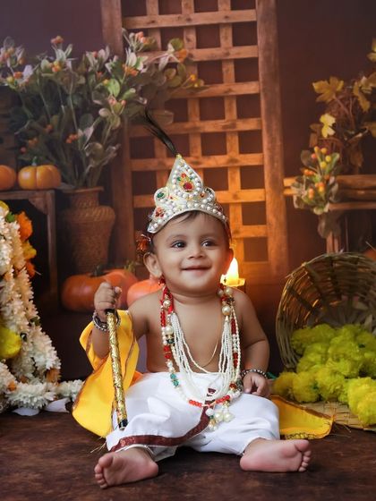 A smiling baby Krishna sits among festive decorations, holding his flute. We aim to capture the innocent and playful nature of Kanha in these special photoshoots.
