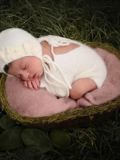 This newborn is curled up in a mossy nest, wearing a white knitted outfit, and placed in a digitally enhanced grassy field for an enchanting look.