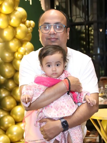 A father holds his baby girl in his arms against a backdrop of golden balloons. A sweet and simple portrait from a family celebration.