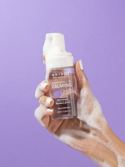 This shot highlights the product's foaming texture. A hand holds the bottle, covered in its own light and airy foam, against a clean purple background that makes the white bubbles pop.