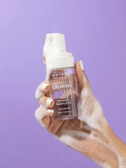 This shot highlights the product's foaming texture. A hand holds the bottle, covered in its own light and airy foam, against a clean purple background that makes the white bubbles pop.
