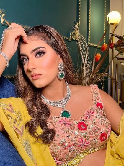A close-up, intense portrait of the model in the floral lehenga, showing the beautiful contrast of the pink blouse, yellow dupatta, and green-stoned necklace.