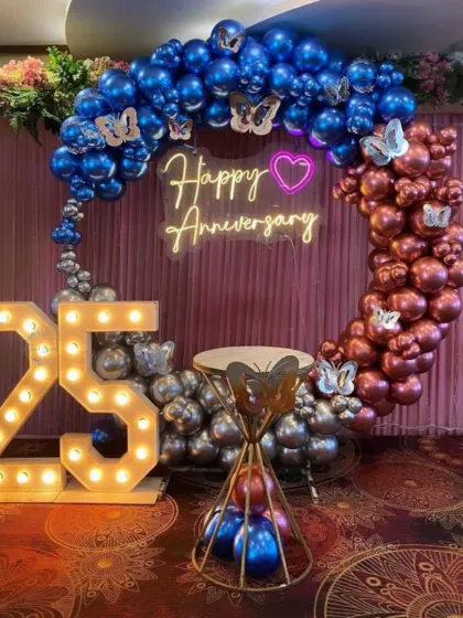 A grand 25th anniversary celebration setup. This decoration features a ring arch with metallic blue, silver, and rose gold balloons, a neon sign, and large light-up numbers.