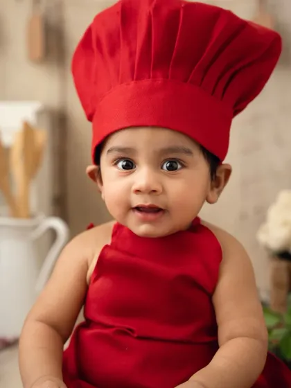 A close-up of a baby dressed as a little chef in a bright red hat and apron. His wide eyes are full of curiosity.