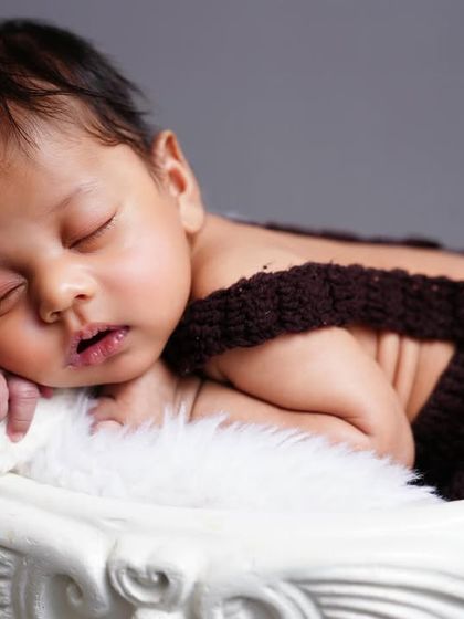 A sweet sleeping newborn posed in a knitted brown outfit, resting peacefully in a white decorative cup prop.
