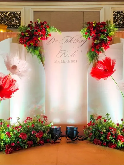A personalized photo backdrop with the couple's name and wedding date, decorated with large, artistic paper flowers and fresh red floral arrangements.