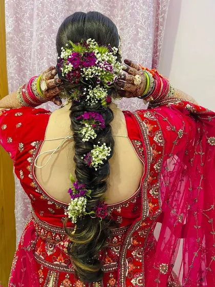 A detailed view of an intricate bridal hairstyle. This fishtail braid is woven with beautiful purple and white fresh flowers, creating a stunning look from the back that complements the bride's red lehenga.